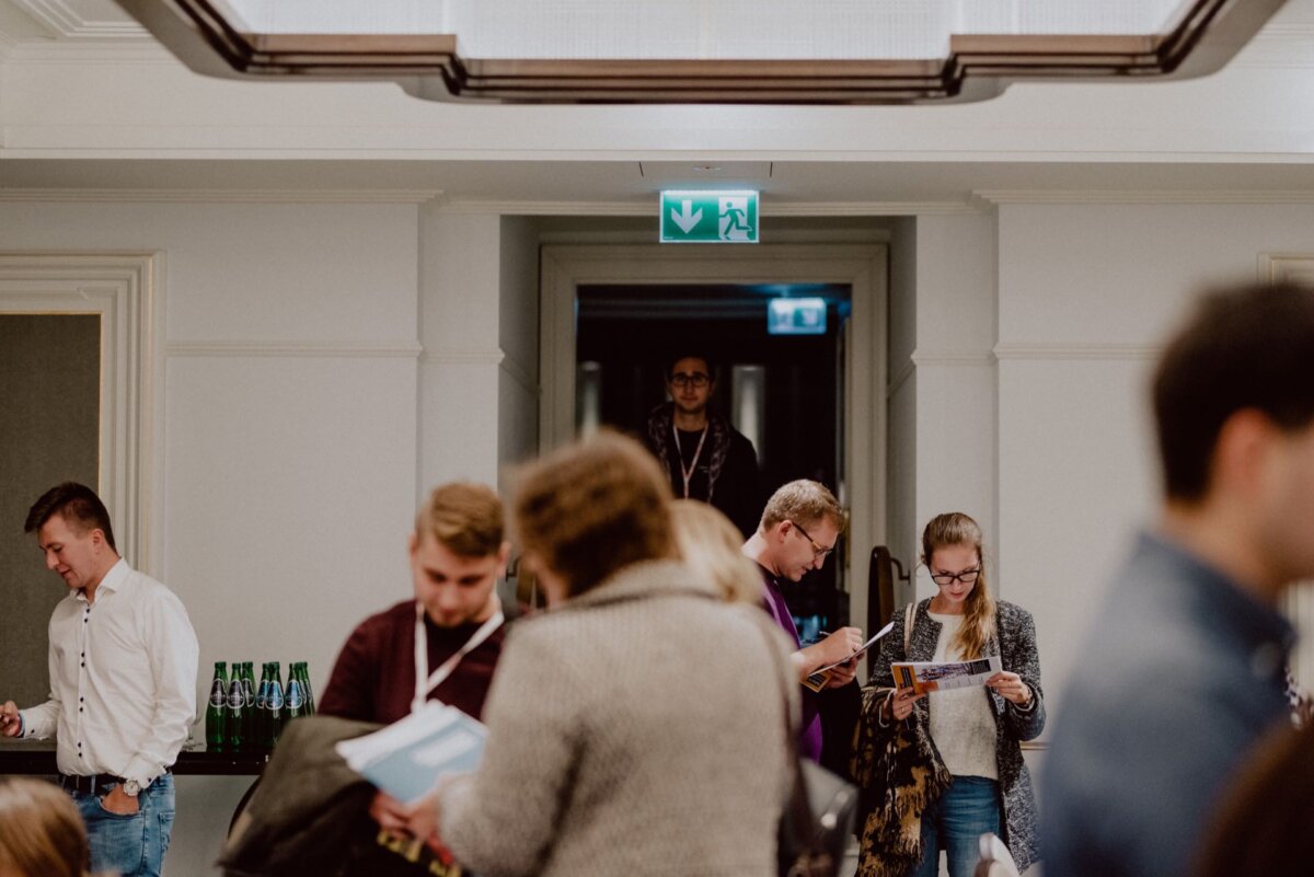 A group of people mingle in a well-lit corridor, ideal for event photography. Some are reading brochures, others are talking. A man stands at the back door, under the illuminated exit sign, and observes the scene. Water bottles stand on a table to the left.   
