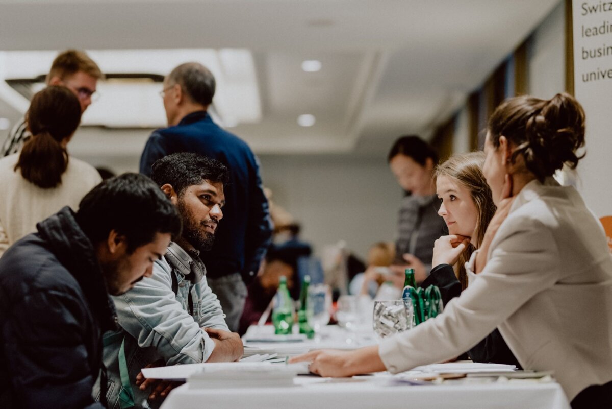 A group of people sits at a table immersed in conversation. Some participants are standing and talking in the background. The scene is reminiscent of a networking event or business meeting, and the professional and tense atmosphere is perfectly captured by *event photography*.  