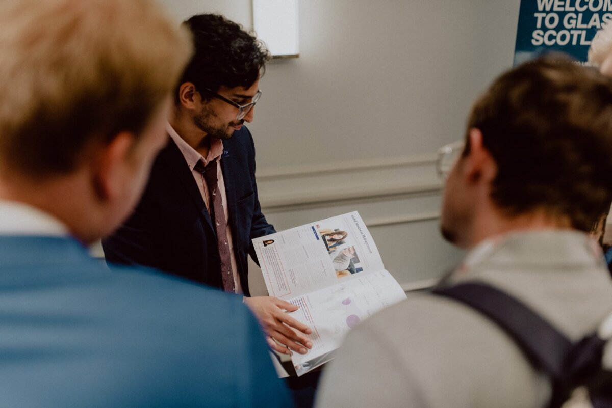 A man in a suit and glasses presents a document or brochure to a small group. The brochure contains text and images. Two people, with their backs turned to the camera, listen intently. A placard in the background proclaims: "Welcome to Glasg (Scotland)." The scene captures the perfect dynamic of event photography.    