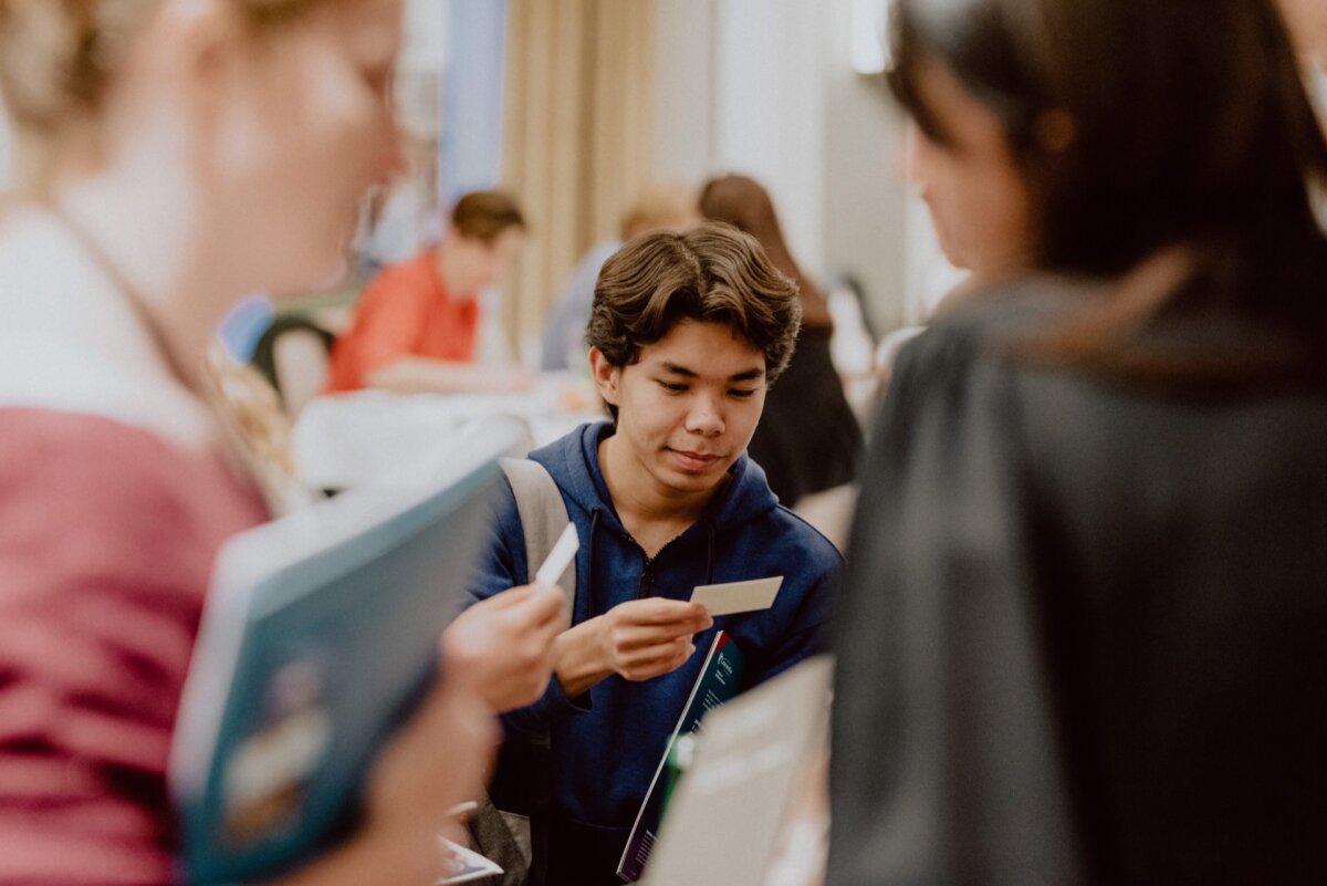 A young person in a blue shirt is reading a card while standing among a group of people in a room. The background is slightly blurred, suggesting a busy or social environment. The photo, perhaps from *event photography*, shows the person's concentration as others nearby look at documents or talk.  