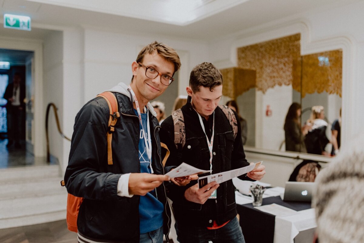 Two young men stand side by side and look at the papers they are holding in their hands. One man, wearing glasses and a backpack, smiles at the camera, while the other, also with a backpack, focuses on reading. They appear to be in the lobby or registration area of the room - perhaps they were captured for a photo essay on the events in Warsaw.  