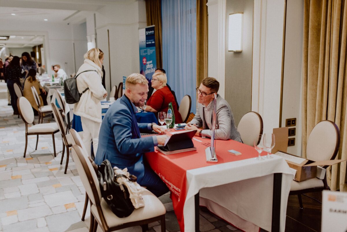 People sitting at tables in a conference room, engaged in discussion. Two men in business attire are sitting at a table with a red tablecloth, one showing the other something on a laptop. Other people can be seen in the background, including a lady in a white apron - perfectly captured by an event photographer Warsaw for event photography.  