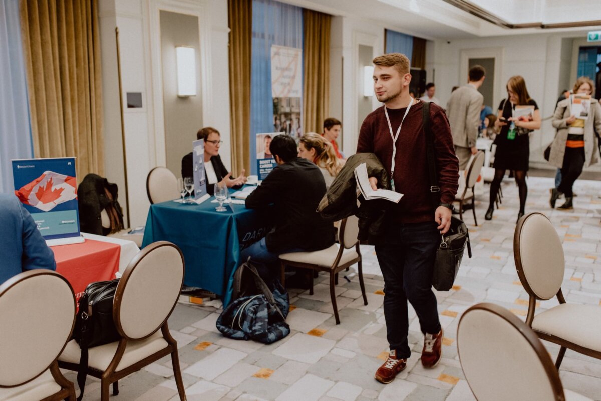 A man walks through the conference room with a lanyard around his neck, holding a coat and a bag. In the background you can see several tables where people are sitting, immersed in discussions. On some of the tables are flags of countries, including the flag of Canada. An ideal scene for *event photography* or *event photography*.   