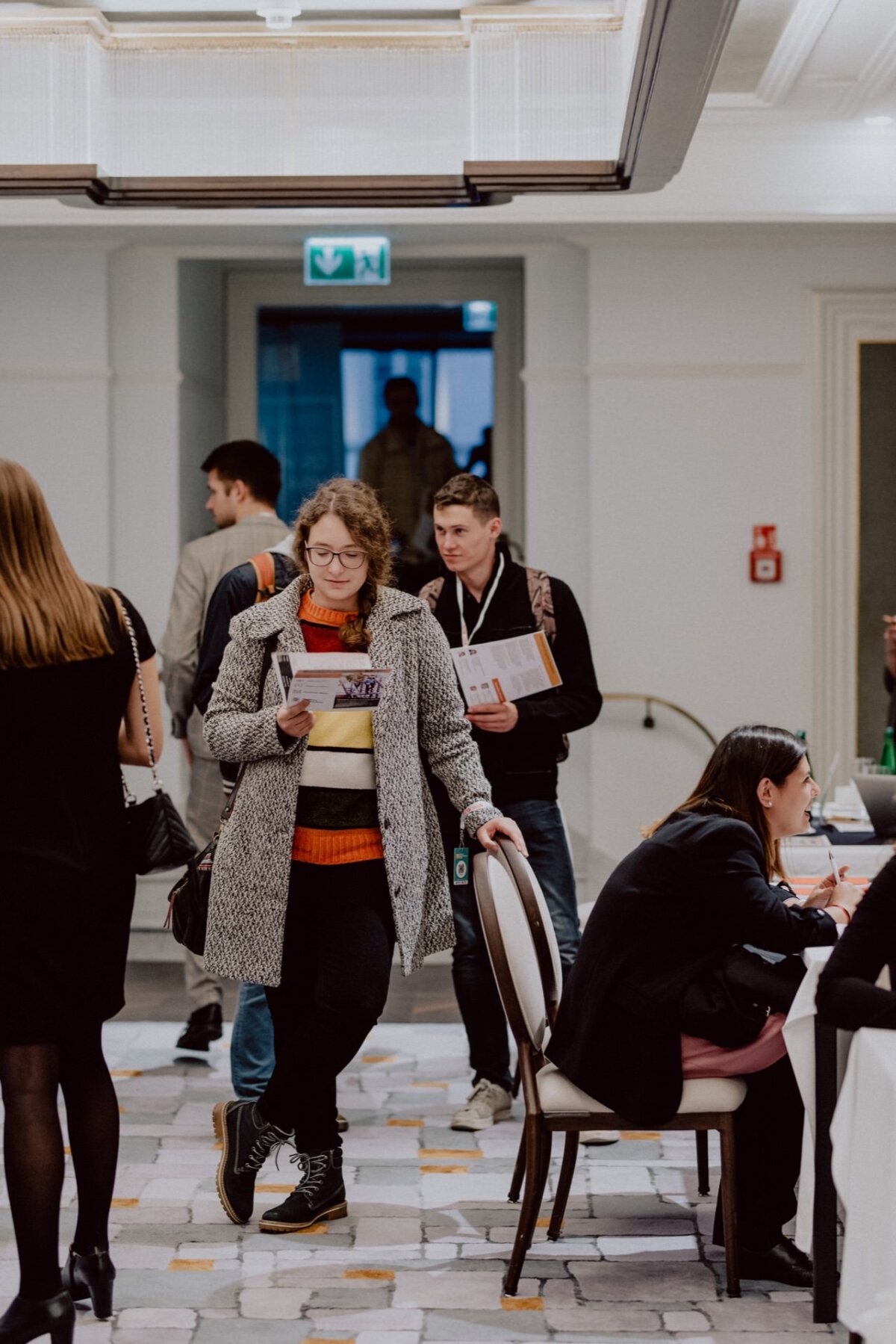 A woman with curly hair, glasses and a coat stands and reads a document in a conference room. You can see people entering the room and doing various activities at the tables. The atmosphere is lively and professional - perfectly captured in this photo report of the event by event photographer Warsaw.  