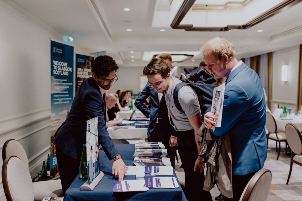 A group of people are standing at a table at a conference or event, listening intently to a man explaining the information on the table. In the background are banners and other tables placed in a well-lit room, perfectly capturing the essence of event photography. 