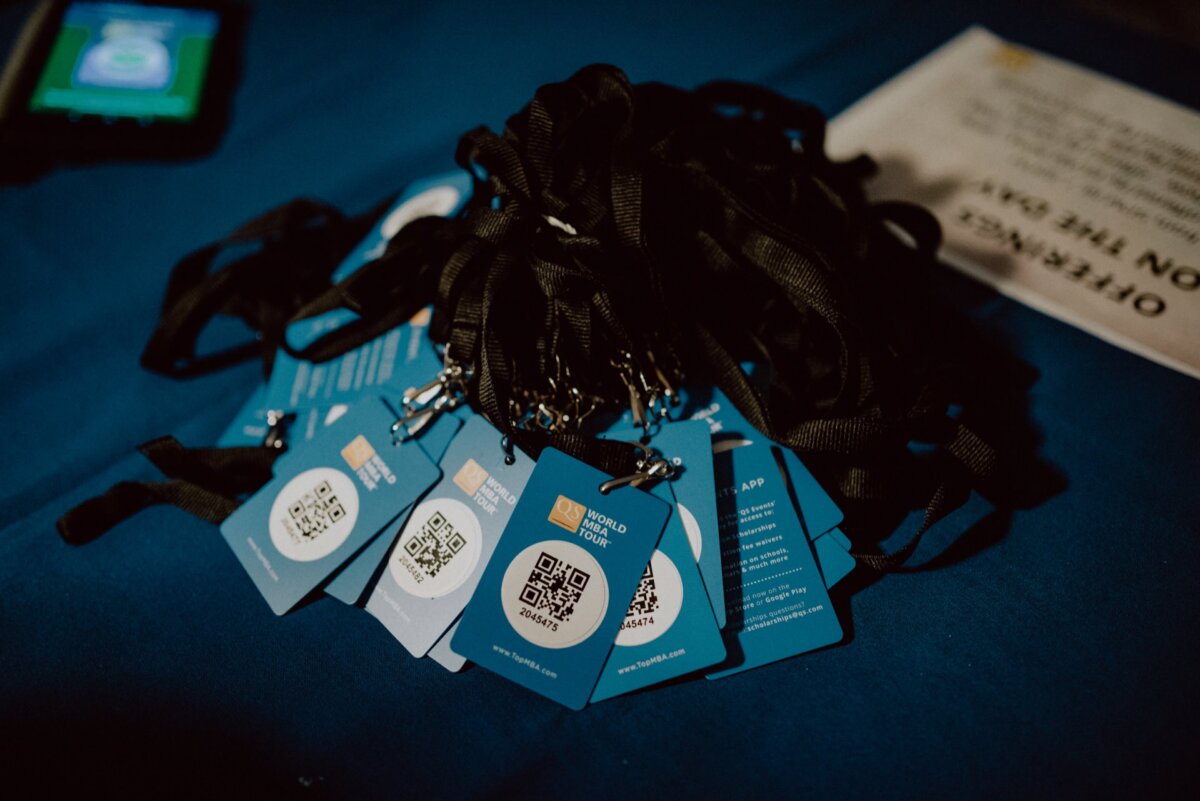 On a blue surface lies a stack of blue conference badges attached to black lanyards, some with visible QR codes. A sign with the words "OFFERS IN APPLICATION" is partially visible in the background. A smartphone is visible near the edge of the photo, capturing a moment of classic event photography.  