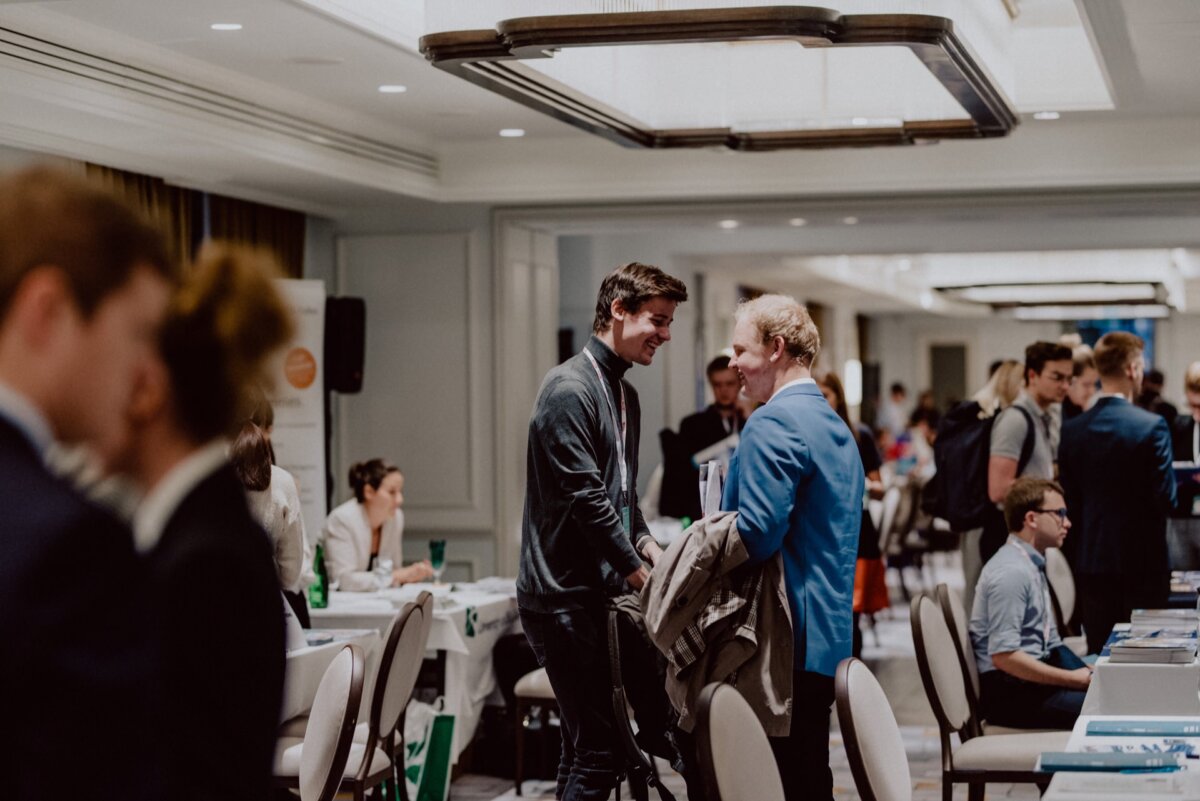 A bustling event in a room with people engaged in conversation. In the middle, two men are talking and shaking hands; one is holding a jacket. Other participants sit or stand and talk at tables. The well-lit room features modern lighting and décor, ideal for photographing events and capturing lively moments.   
