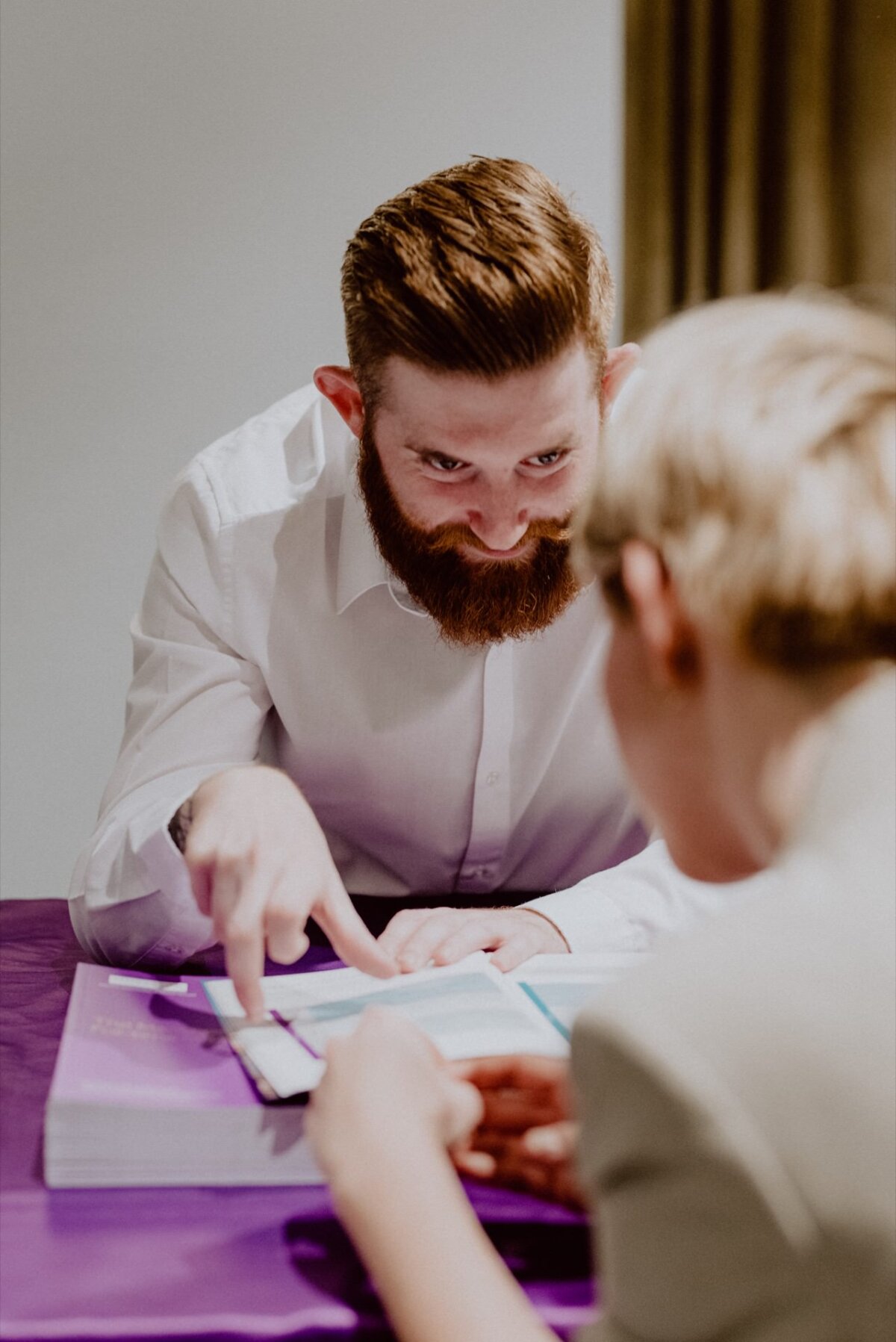 A man with a brown beard points to a tablet screen, talking and smiling at a person with short blond hair sitting across from him. They sit at a table with purple books and a purple tablecloth, illustrating moments captured in an event photograph. 