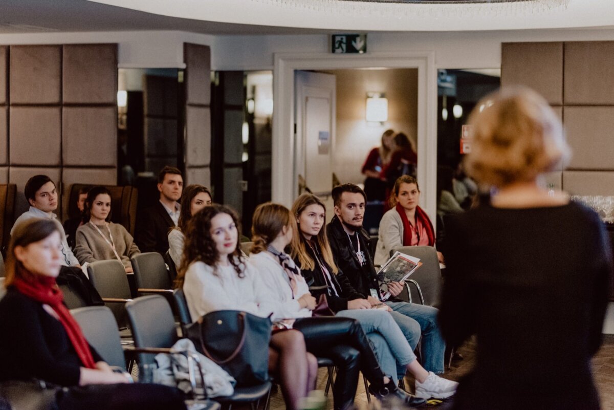 In a dimly lit room, a group of people sit in rows of chairs and listen intently to the speaker, whose back is turned to the camera. The formal atmosphere underscores the engaging presentation, perfect for an event photo shoot or event photography. 