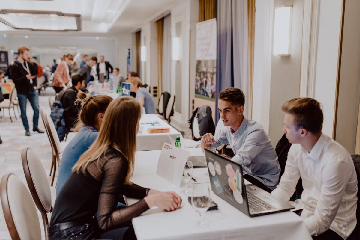 A group of people, mostly young adults, are having a conversation at a networking event or conference in a spacious, well-lit room. They are seated at round tables with laptops and business cards, framed ideally with photo coverage of the events. Other participants and tables are visible in the background.  