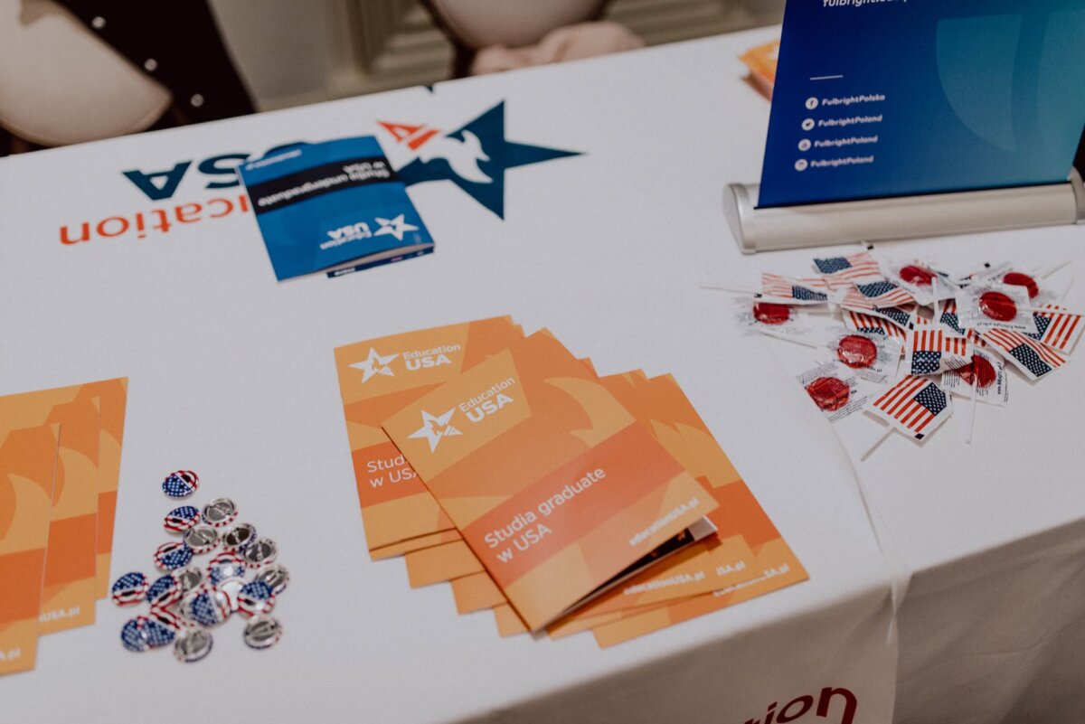 A display table with orange brochures titled "Studying and Graduating in the USA," pins with American flag motifs and various red lollipops in American flag wrappers. A tablecloth and vertical sign display the "EducationUSA" branding, perfect for capturing in a photo recap of the event. 