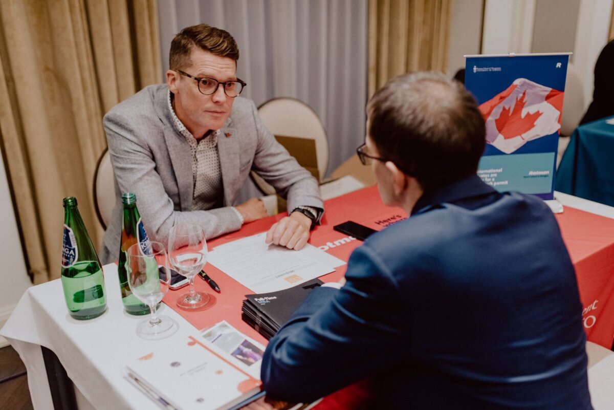 Two men sitting at a table covered with a red tablecloth are talking. One, with short hair and glasses, wears a light gray jacket; the other, with short dark hair and dark glasses, is wearing a navy blue suit. On the table lie documents, water bottles and signs of a Canadian brand - an excellent photo-record of events.  