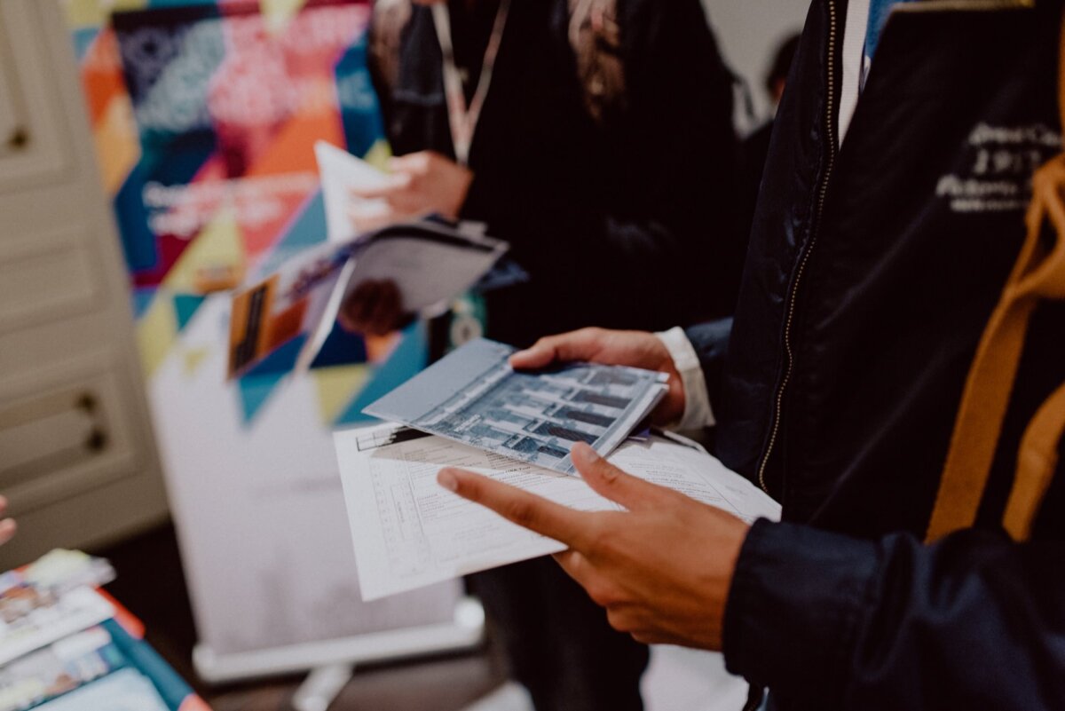 Two people are standing and reading documents or pamphlets. The hands of one person holding the papers are visible, while the other person holds the document close to his chest. In the background is a colorful presentation board that captures the essence of event photography from a recent event photo essay.  