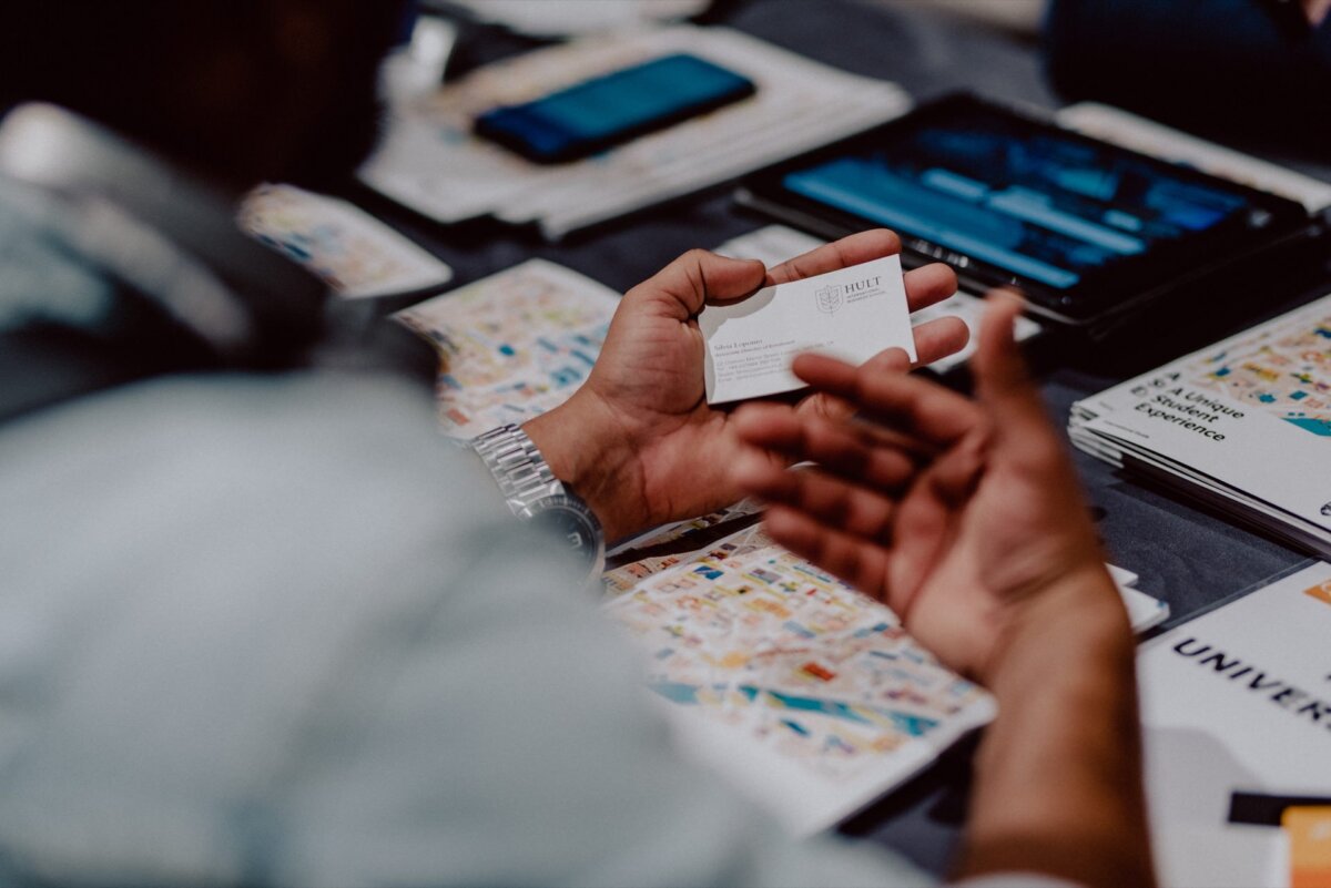 A person holding a business card with both hands, surrounded by various documents and a tablet on a table. The documents include colorful charts and brochures. A blurry figure can be seen in the foreground, focusing on the material held in his hands - capturing every detail for event photography.  