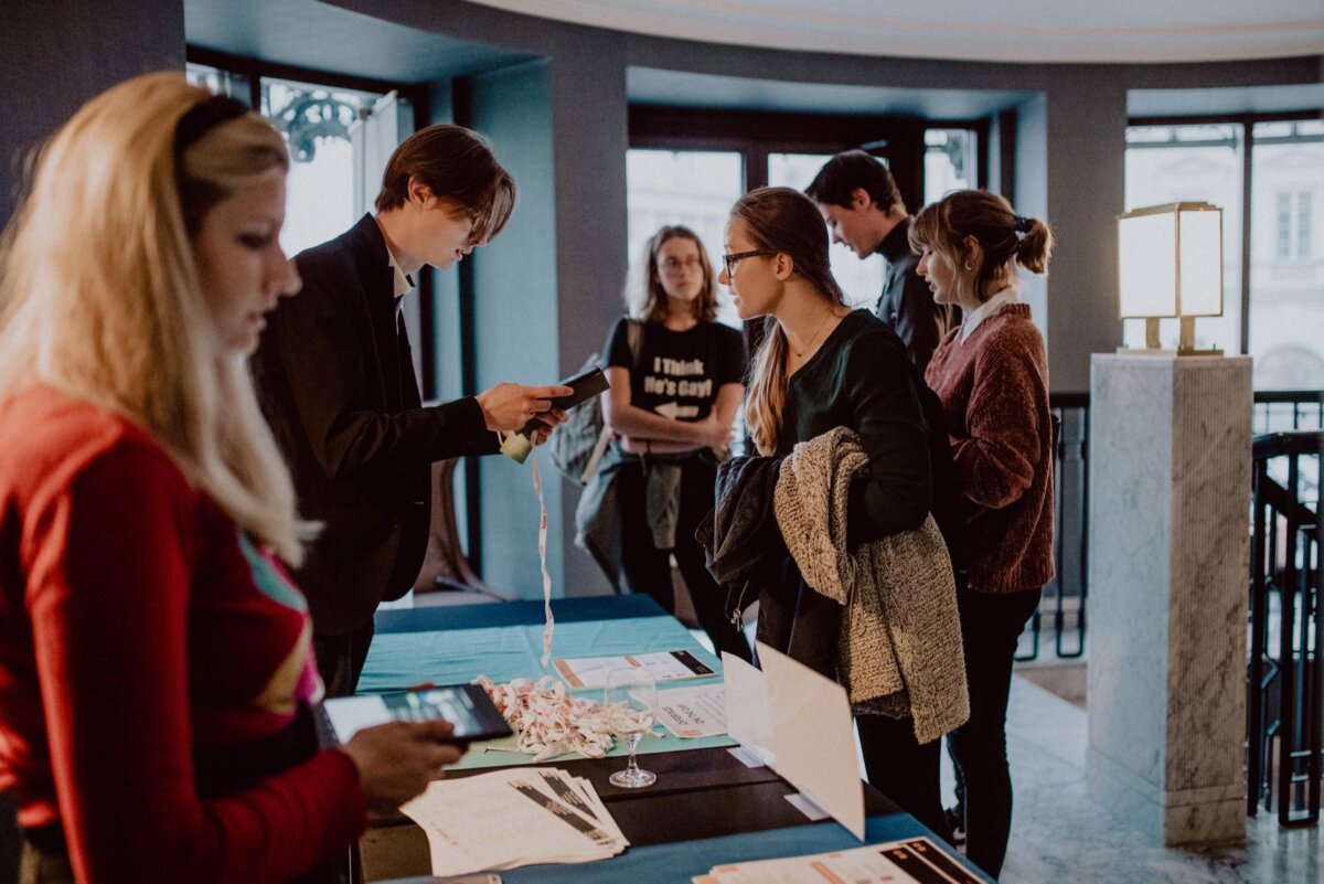 People are gathered around a table of informational materials in a modern, indoor setting. It looks like a few people are logging in or registering for an event, while others are busy chatting under a well-lit, contemporary design. This photo report of the events perfectly captures the vibrant atmosphere.  