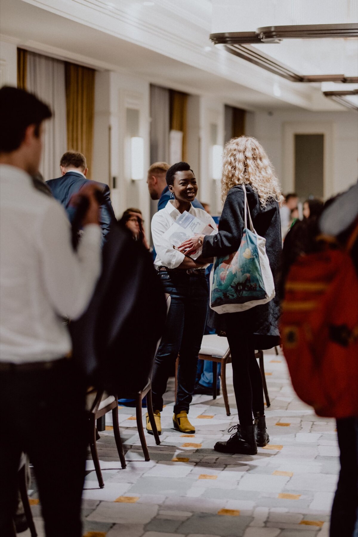 Two people greet each other in a busy, well-lit corridor. One person is wearing a white shirt and yellow shoes, the other has curly blond hair and carries a large bag. In this action, this moment captures the essence of event photography. Other people can be seen moving in the background.   