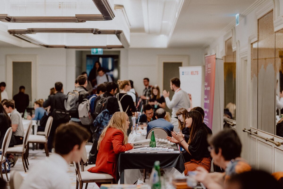 The busy, modern conference room is filled with people having conversations. Some sit at tables and talk, others stand in groups. The room has a warm atmosphere, bright lighting and modern decor. Large banners and materials are visible in the back; perfect for an event photo shoot.   