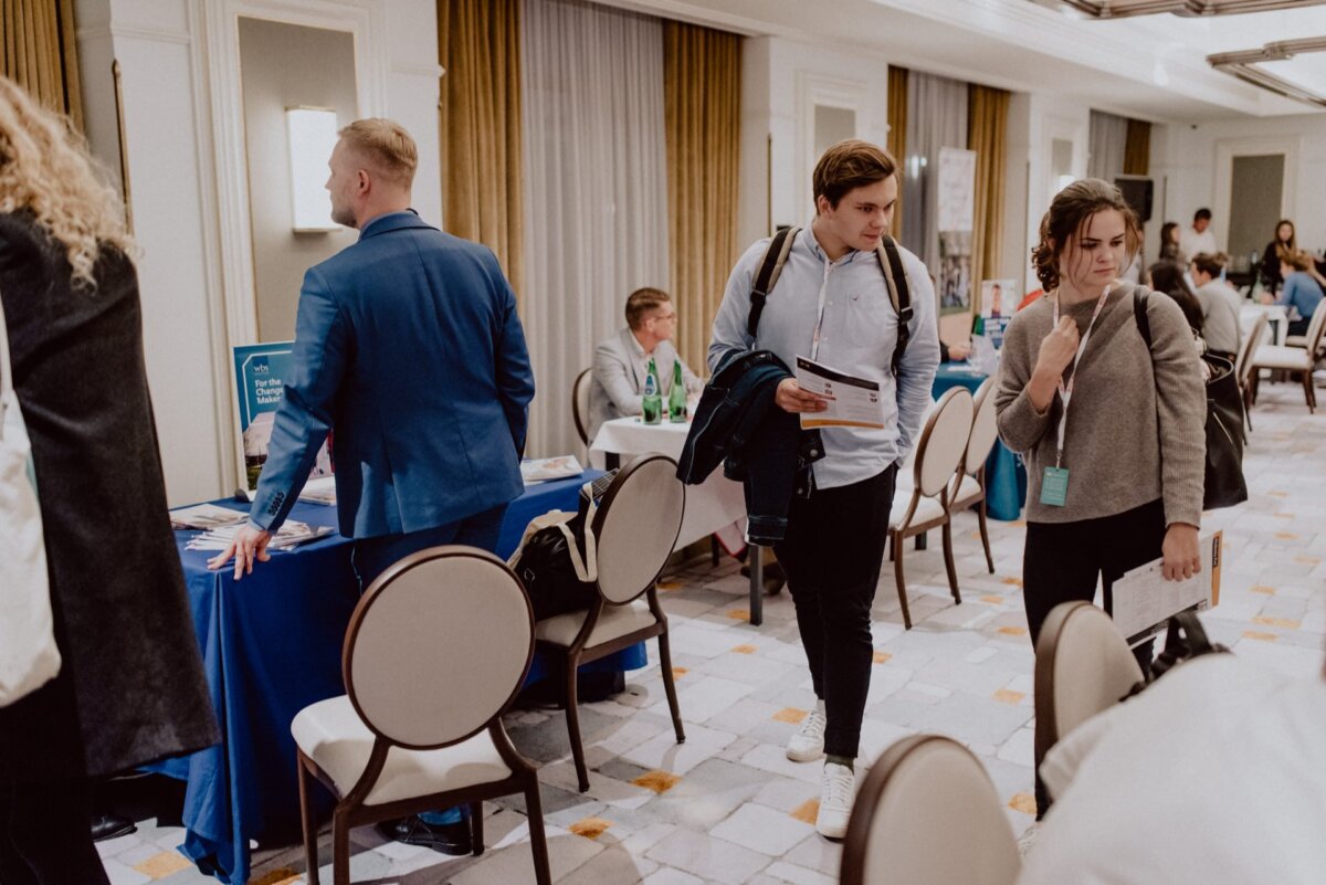 At a professional event in a conference room, people discuss at tables while others walk around. Tables are adorned with office chairs and handouts. Event photography captures these interactions, highlighting the dynamics and atmosphere, perfect for event photojournalism.  