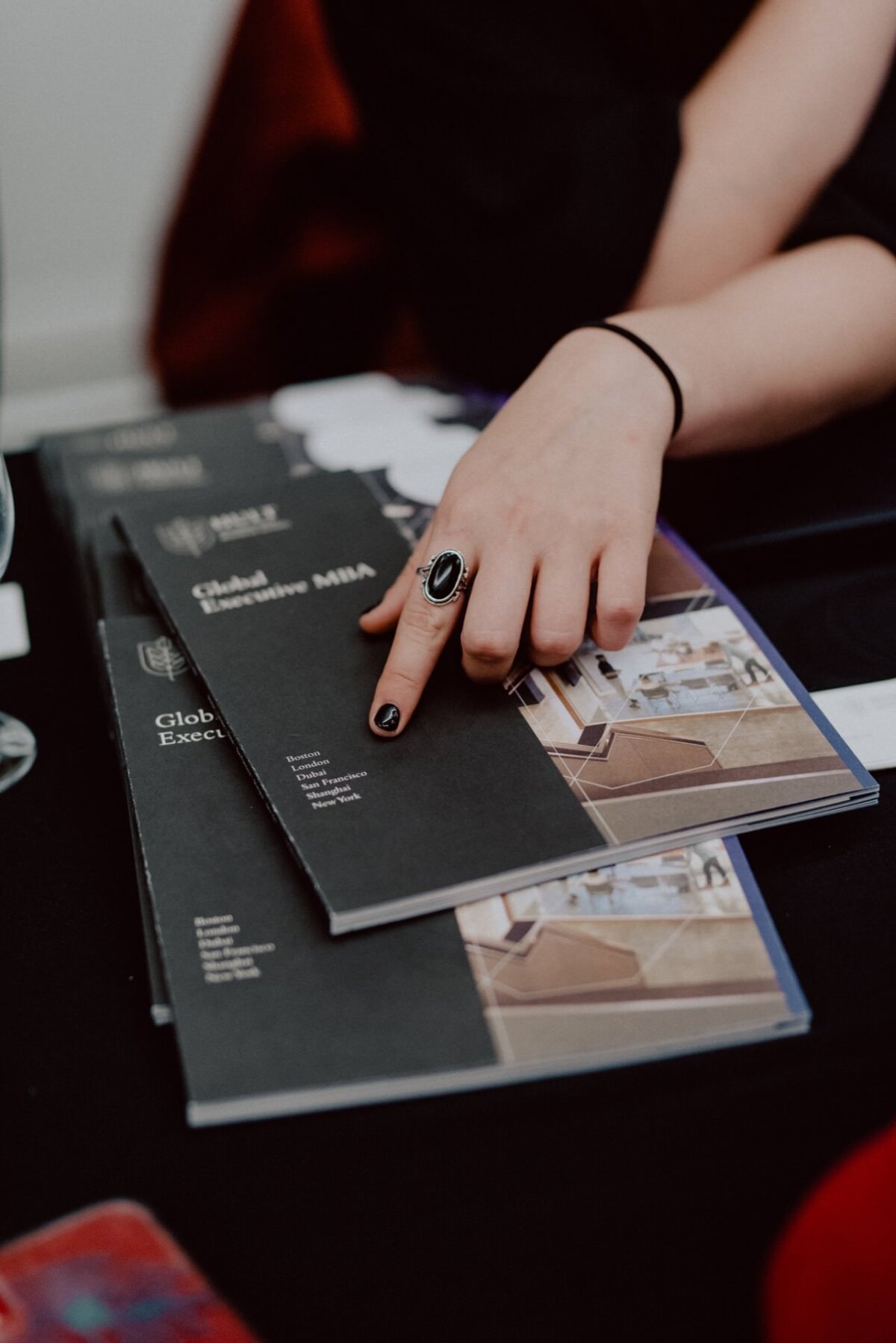 A person with a black ring on his finger points to a brochure lying on the table titled "Global Executive MBA." More brochures containing photos of modern interiors are stacked underneath. The person's hand rests on the table, capturing a moment worthy of an event photographer Warsaw.  
