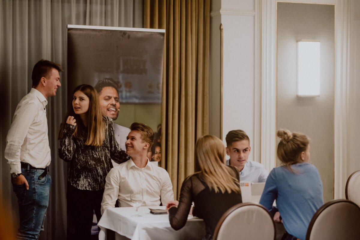 A group of young people gather in a well-lit room. Some are sitting at a table, talking and smiling, others are standing nearby. A banner with a smiling face is partially visible in the background. The atmosphere seems friendly and relaxed, perfect for an event photographer Warsaw to capture every moment.   