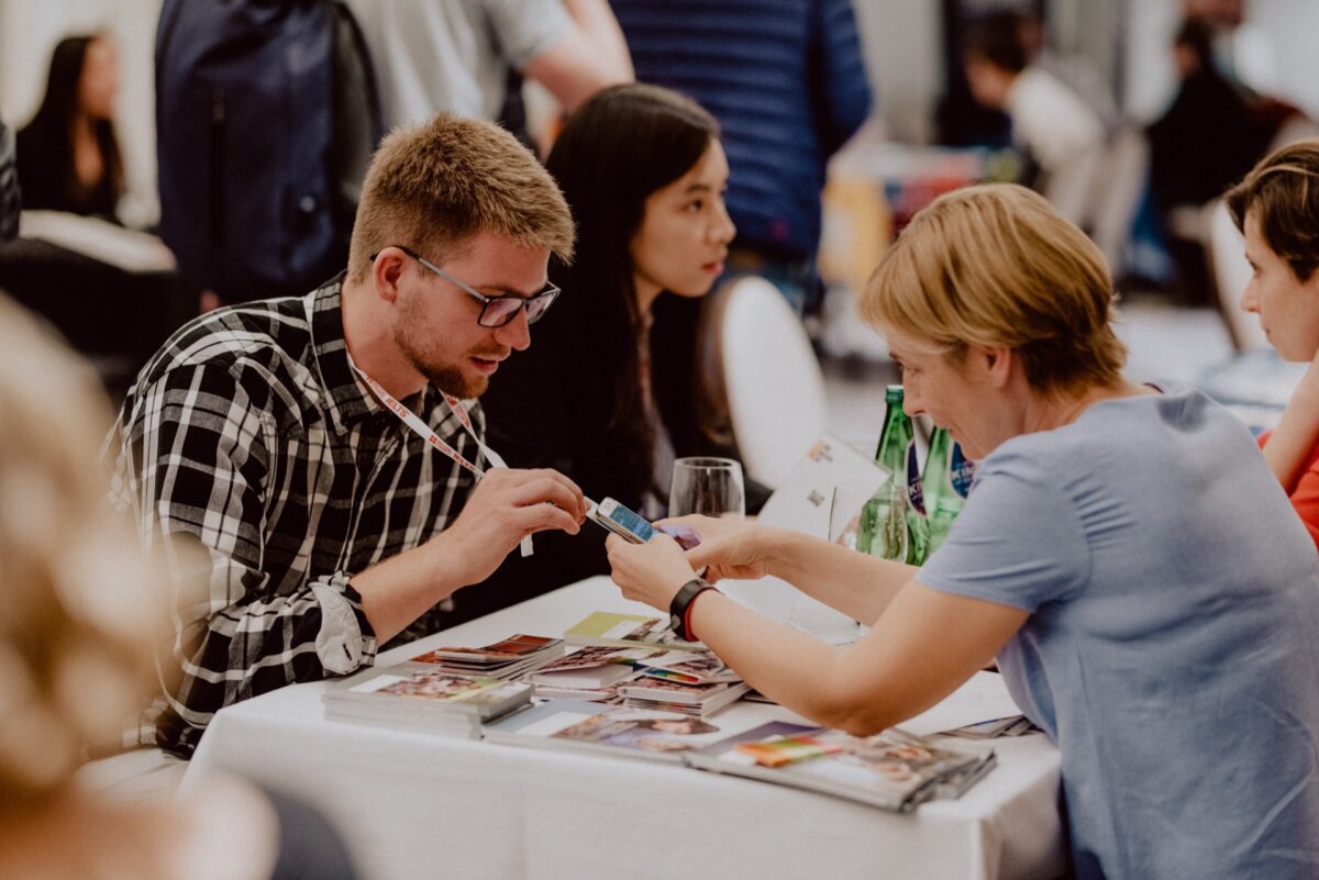 There are several people sitting at a table having a conversation and looking through various documents and brochures. A man in a checkered shirt and glasses is talking to a woman in a light blue shirt, who is showing him something on her phone - capturing the essence of event photography. 