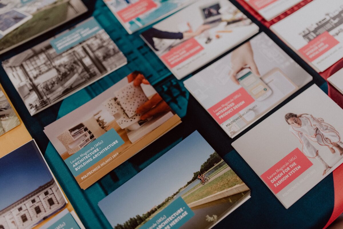 A table covered with a variety of cards and brochures showcasing various architectural and design projects, captured in a photo essay of the events. Each card contains photos, rendered images and descriptions of various concepts and projects, covering a mix of buildings, interiors and product designs. 