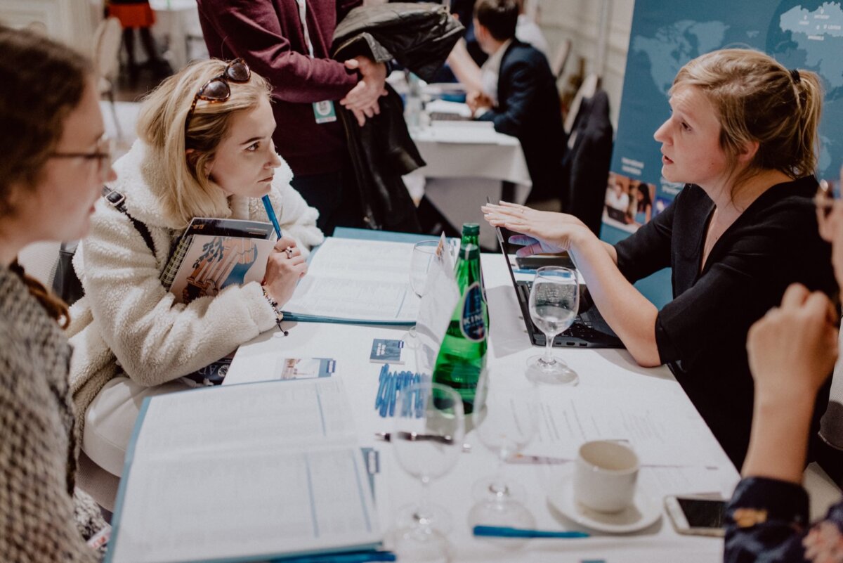 Three women are having a conversation at a conference table. One woman on the right is talking and gesturing expressively, the other two are listening attentively. On the table lie documents, a laptop, a bottle of water and a glass. Other participants are in the background. This scene perfectly captures the essence of event photography.    