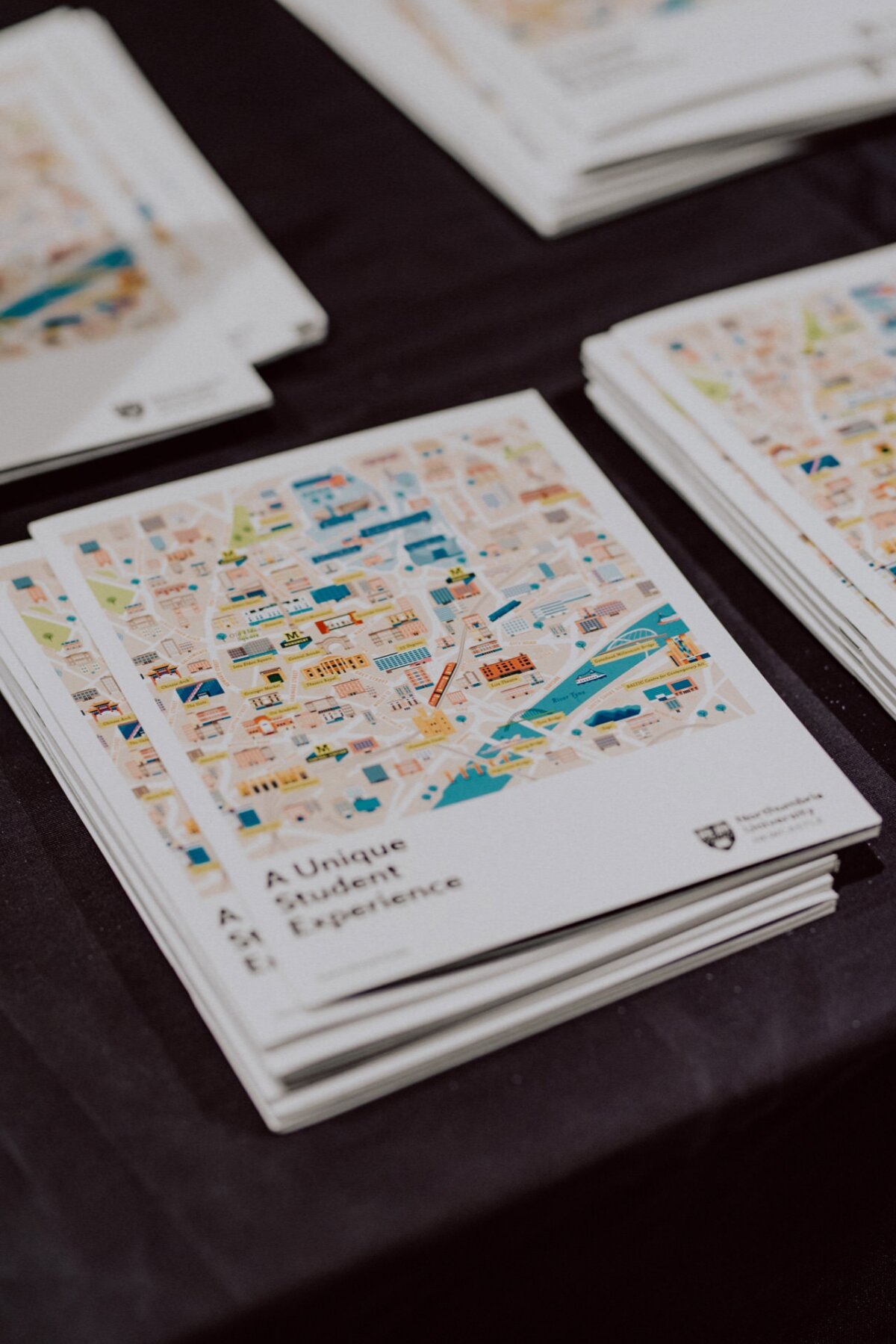 Stacks of brochures titled "Exceptional Student Experience" with color illustrations showing the layout of the campus displayed on a black tablecloth, beautifully captured in a photo recap of the events. The brochures include a detailed map showing various landmarks and locations. 