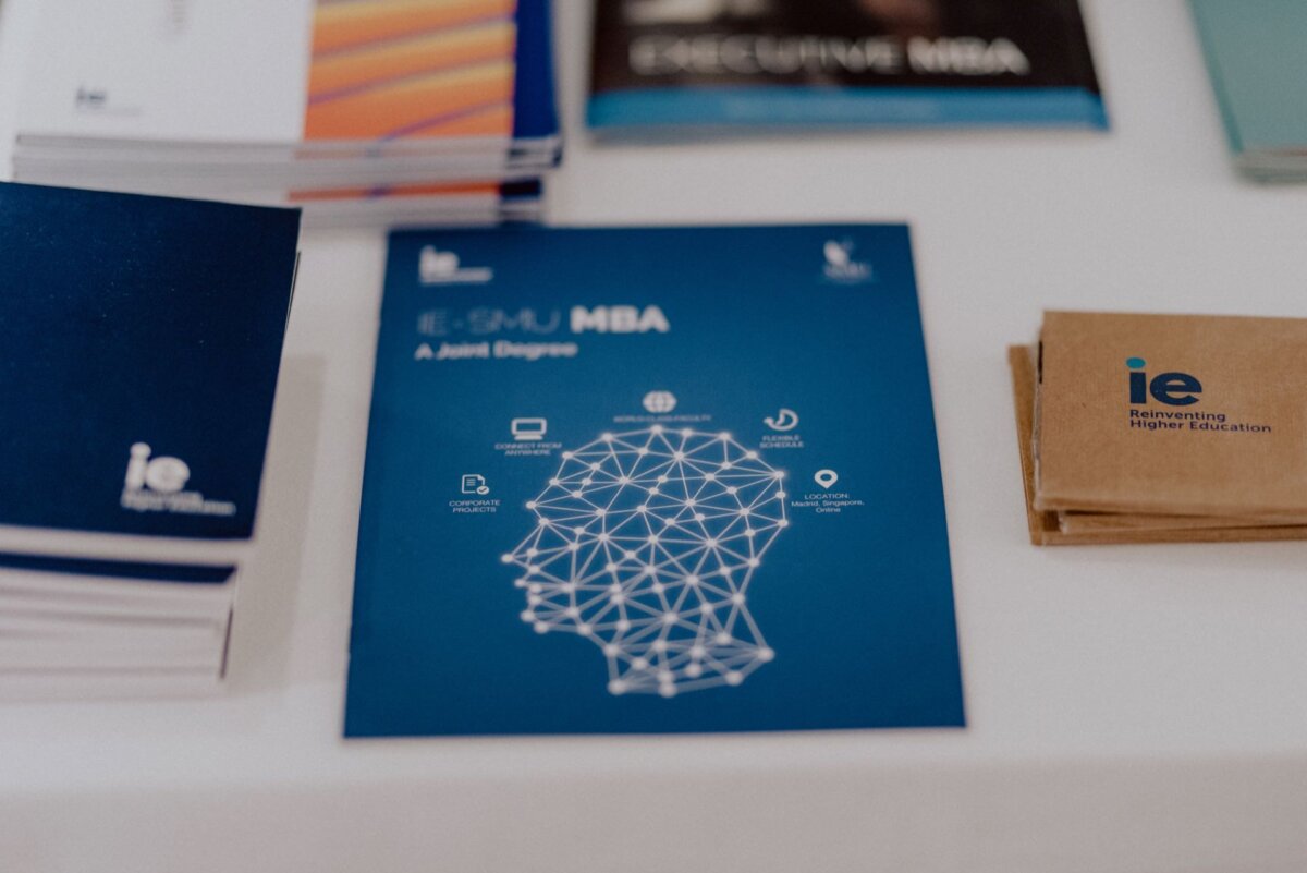 Close-up of brochures and business cards on the table. The central brochure is titled "IE-SMU MBA" and features a geometric design of a human head with icons of various concepts related to education. The business cards bear the IE logo and the slogan "Rediscovering Higher Education." This scene captures the essence of event photographer warszawa, presenting carefully designed materials during an educational event.   