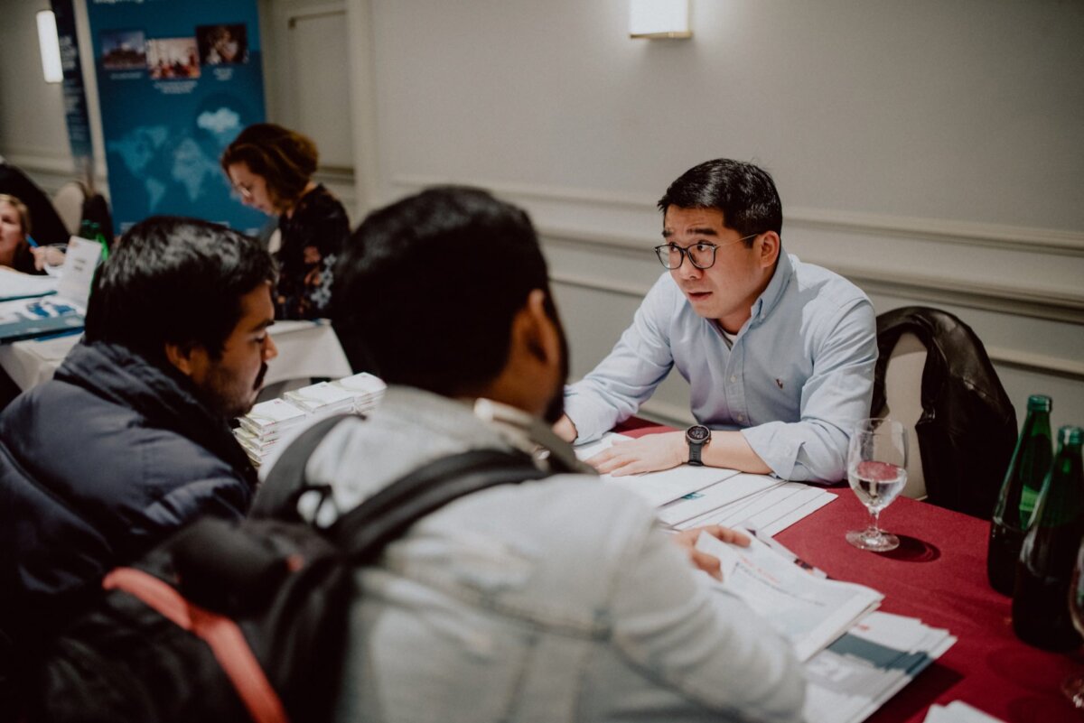 A man with glasses is sitting at a table in a conference room, talking with two other men across from him. Papers are spread out on the table, and a glass of water and bottles stand nearby. Other people can be seen in the background, also in discussion - perfect scenes for event photography.  