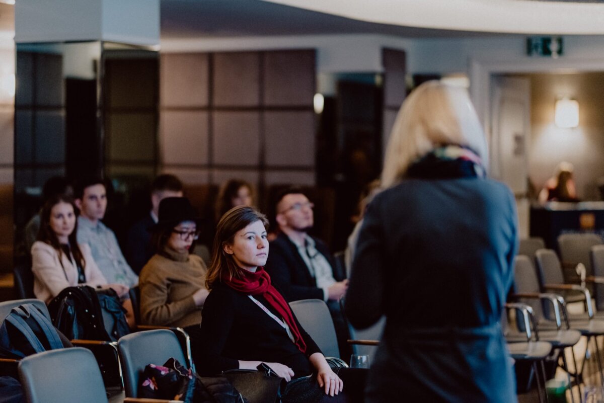 A group of people sit on chairs and listen attentively to a speaker at an event taking place in the room. The blond-haired speaker is partially visible in the foreground from behind. The audience is focused on the presentation, some taking notes. This scene captures the essence of professional event photography.   

