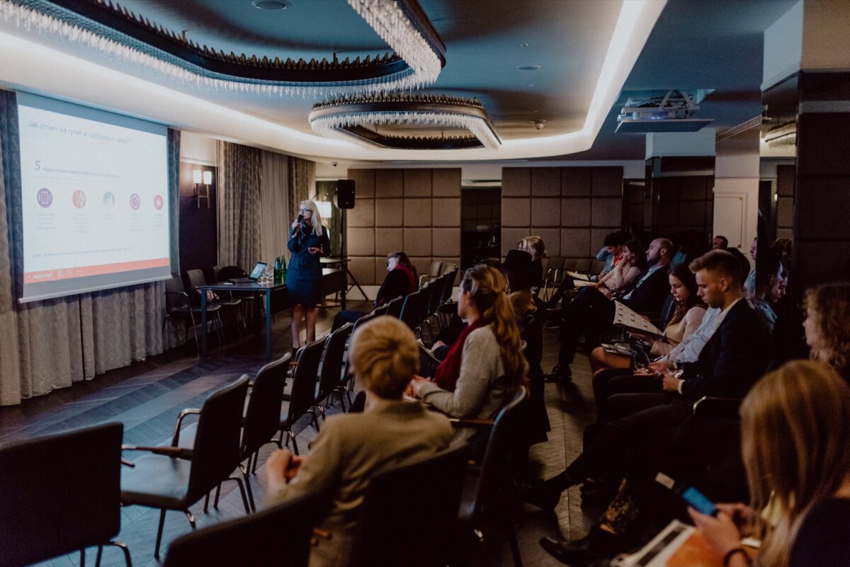 A group of people sit in rows of chairs facing a presenter in a conference room. The presenter stands near a projector screen and displays a slide of the presentation. The room is dimly lit, with ornate ceilings and modern interior design, perfect for capturing photo coverage of events by event photographer Warsaw.  