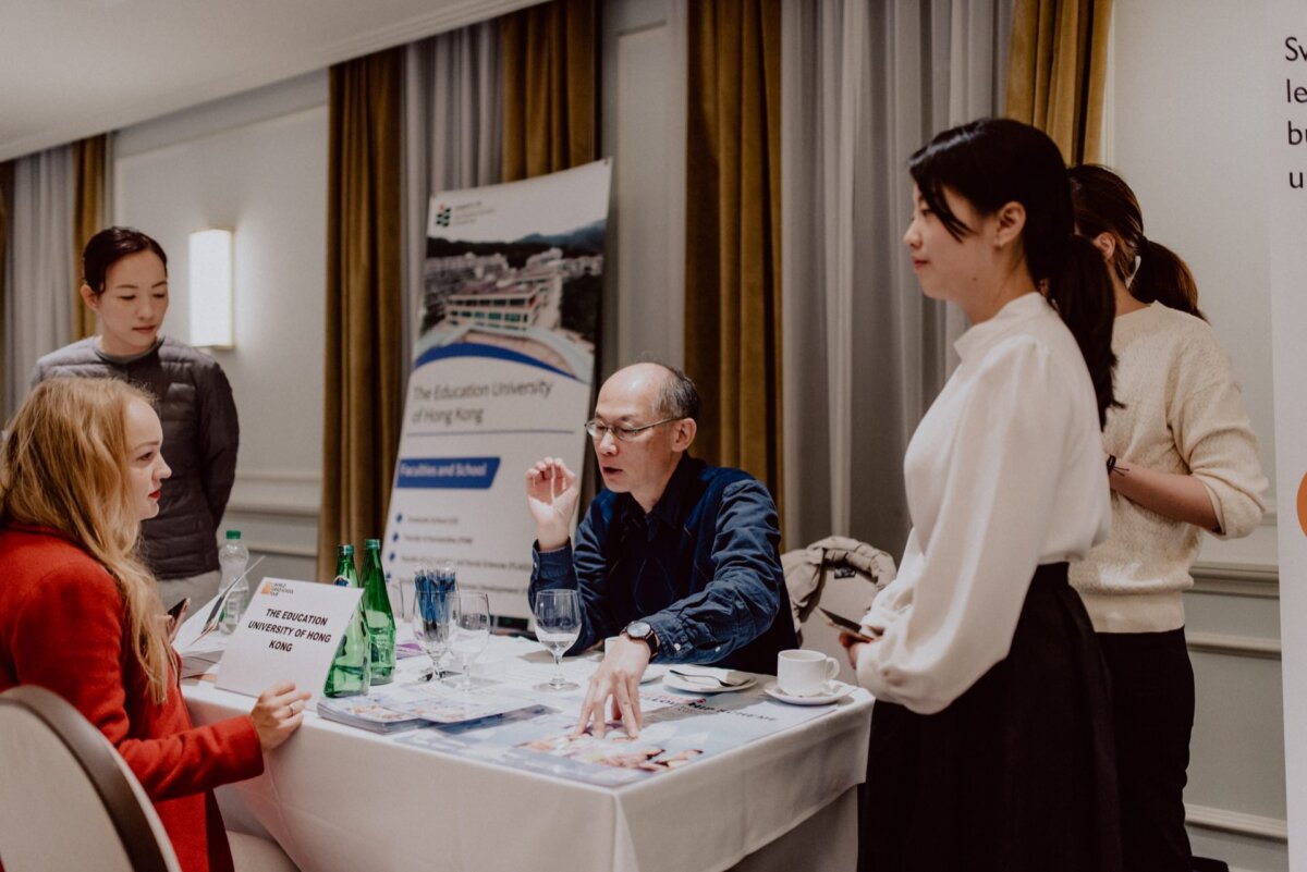 A group of people participating in a discussion at an event held in a room. A man sitting behind a table with a placard reading "Hong Kong University of Education" is talking to a woman and three other people standing around him. There are banners and water bottles on the table, capturing the essence of the photojournalism of the events.  

