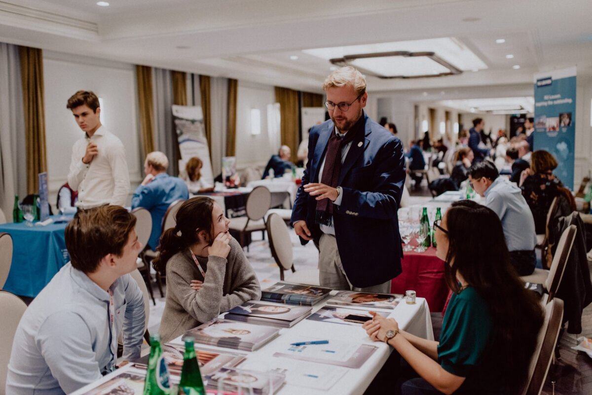 A man in a suit is standing and talking to a group of people sitting around a conference table covered with papers and water bottles. In the background, more people are discussing at various tables in a large, well-lit room. This scene captures the essence of *event photography* at its best.  