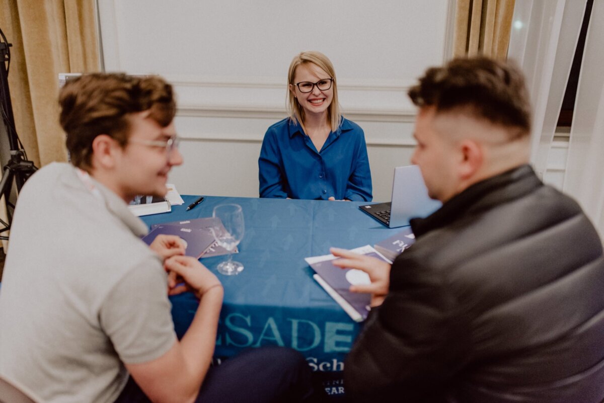 Three people are gathered around a table covered with a blue tablecloth. A woman in a blue shirt and glasses sits to one side, smiles and looks at the two men sitting across from her. The men are immersed in conversation, with papers and a glass on the table, creating the perfect scene for event photography.  