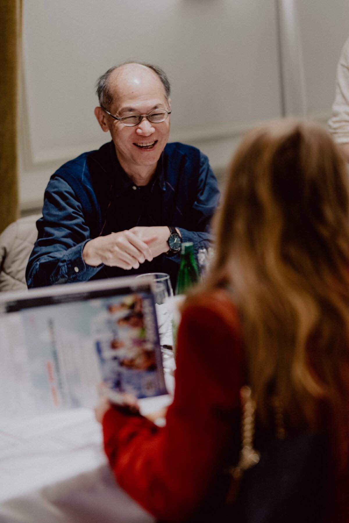 A person wearing glasses smiles and points while sitting at a table during a meeting or meal. Opposite them, another person with long hair holds a document or magazine. The setting seems casual and indoor - perfect for event photography to capture authentic interactions.  