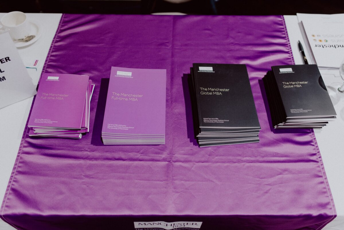 Stacks of brochures titled "The Manchester Full-time MBA" and "The Manchester Global MBA" are stacked on a purple tablecloth. The brochures, with purple and black covers respectively, catch the eye. In the background, a partially visible signboard and documents suggesting a detailed photo recap of the events.  