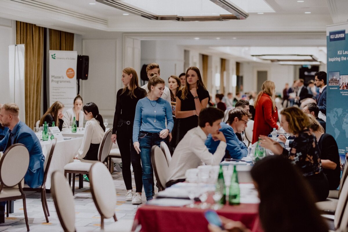 A busy room with many people discussing at various tables, some reviewing documents and others talking. In the foreground, several people are standing in line waiting their turn. Banners and promotional materials are visible in the background - an ideal scene for event or event photography.  