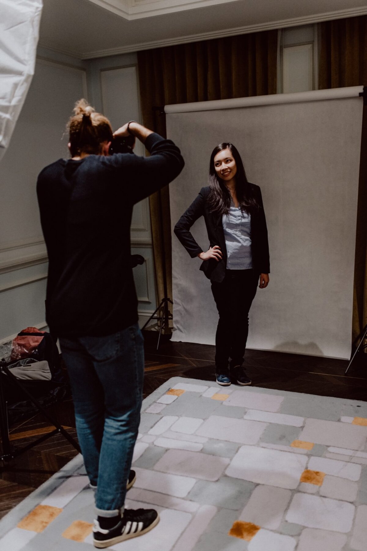 A photographer wearing jeans and a black shirt takes a picture of a smiling woman with long dark hair. The woman, holding one hand on her hip, stands on a tiled floor against a white background. In the background you can see the event photographer's equipment.  