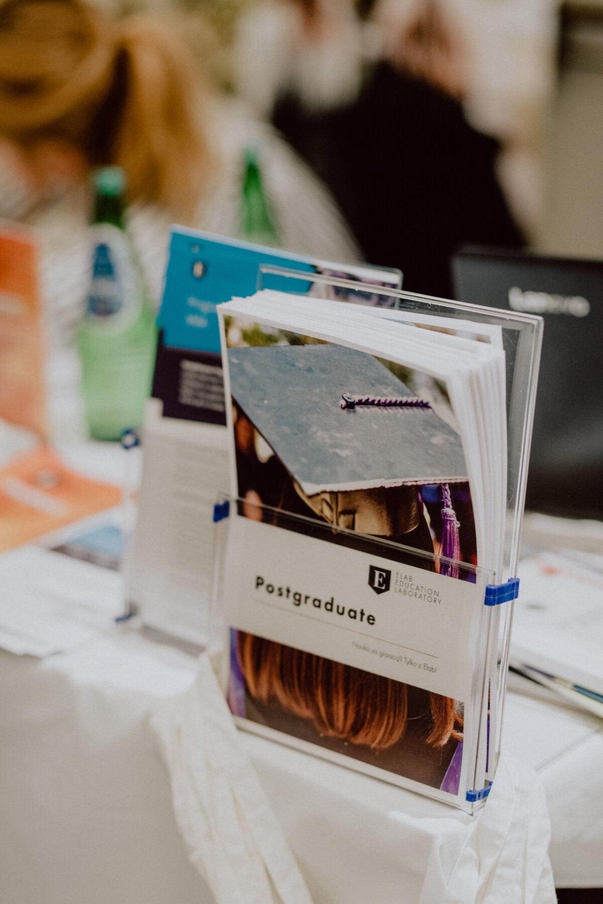 There are several brochures on the rack, including one titled "Postgraduate" with an image of graduates in caps and aprons. The brochures are neatly arranged on a table next to a green glass water bottle and other marketing materials, perfect for those interested in event photography. 