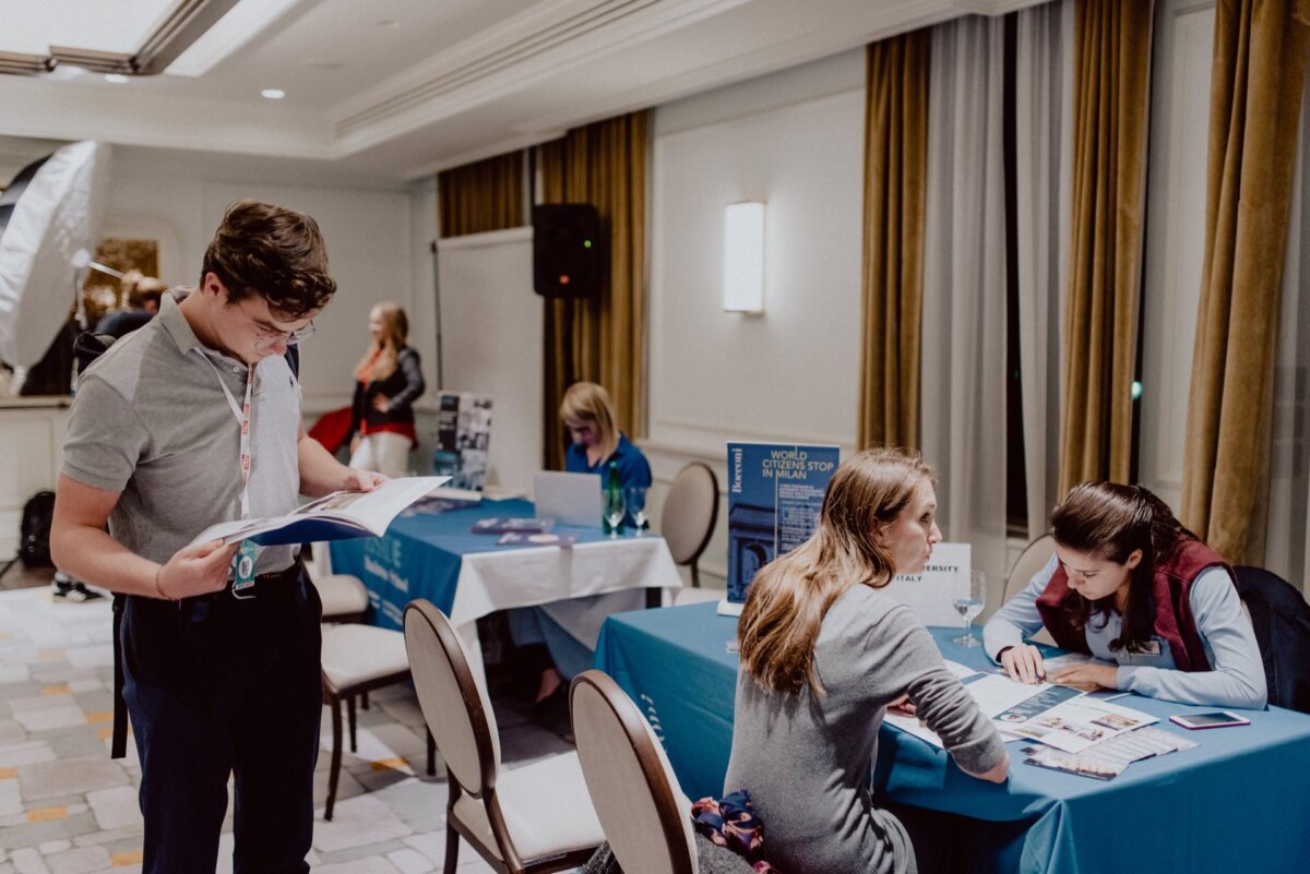 A man reads a brochure while two women sit at a table and talk. Behind them, a third woman sits at another table and works on a laptop. The room captured in this photo essay of the event has tables covered with blue tablecloths, white walls and large windows with yellow curtains.  