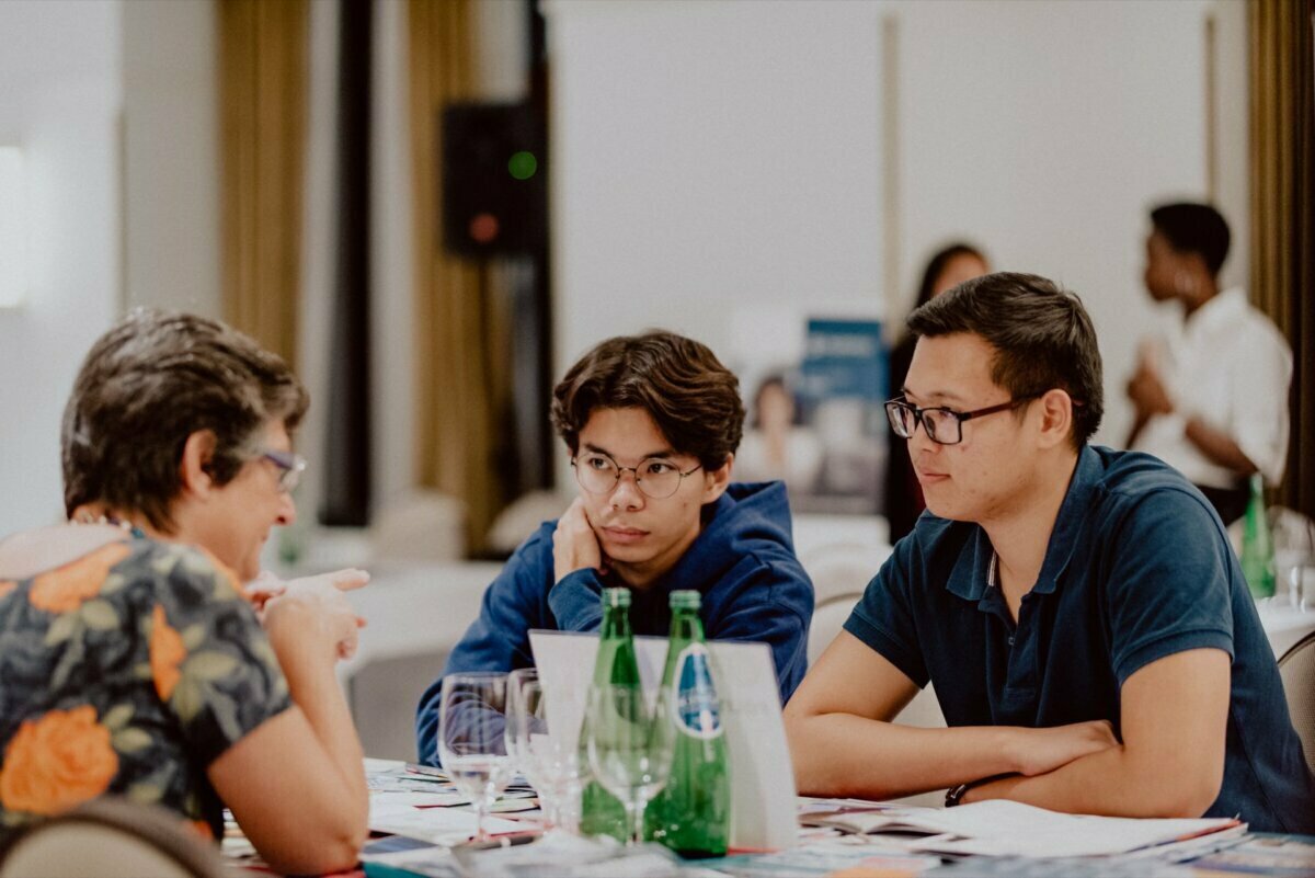 Three people sit at a table engrossed in discussion. The two on the right are listening intently to the speaker on the left, surrounded by water bottles, glasses and papers. Other people can be seen in the background, capturing the perfect moment for *event photography* or *event photography*.  