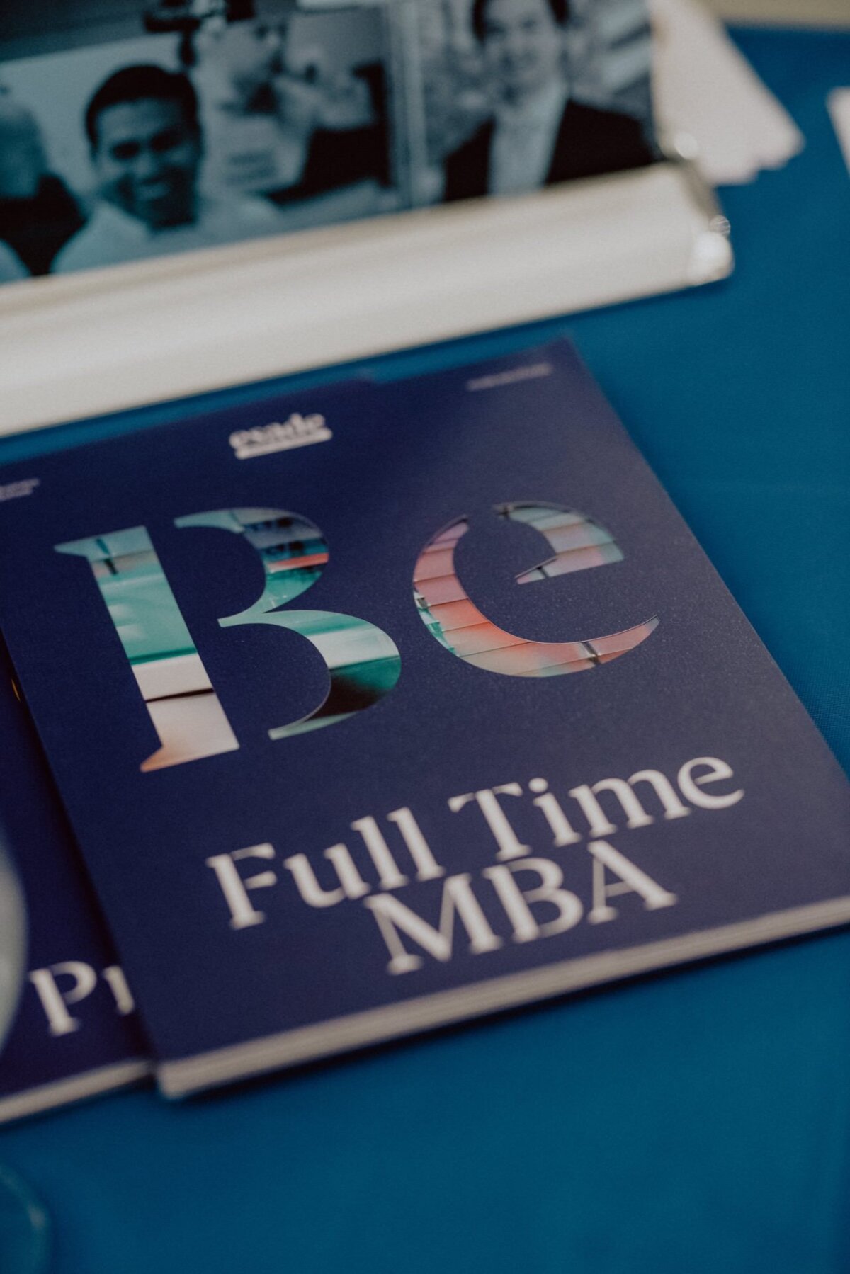 A brochure titled "Be Full Time MBA" lies on a blue tablecloth. The word "Be" appears in a colorful, reflective pattern. The partially visible documents and blurred background of the photo event add depth to the scene, reminiscent of event photography materials.  