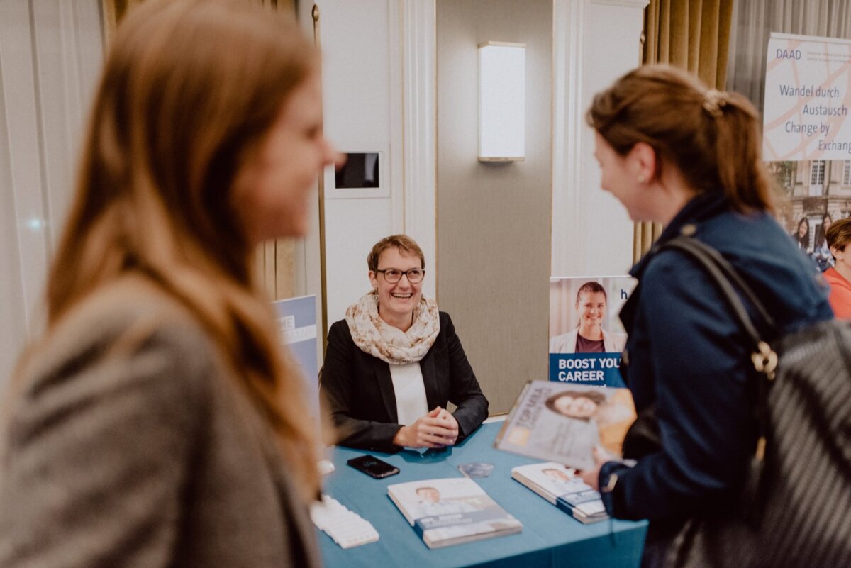 People are interacting at a career fair booth. A woman sitting at the booth smiles and hands out materials, while two other women stand and interact with her. The booth has a sign that reads "Develop Your Career" and some informational flyers on a table - perfect timing for event photography.  