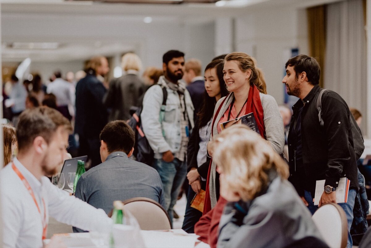 A group of people are standing and talking in a busy conference room. Some are holding books or notebooks, others are sitting at tables, immersed in discussions. The atmosphere is lively and conducive to collaboration, perfect for an event photo report taken by a skilled event photographer warsaw.  