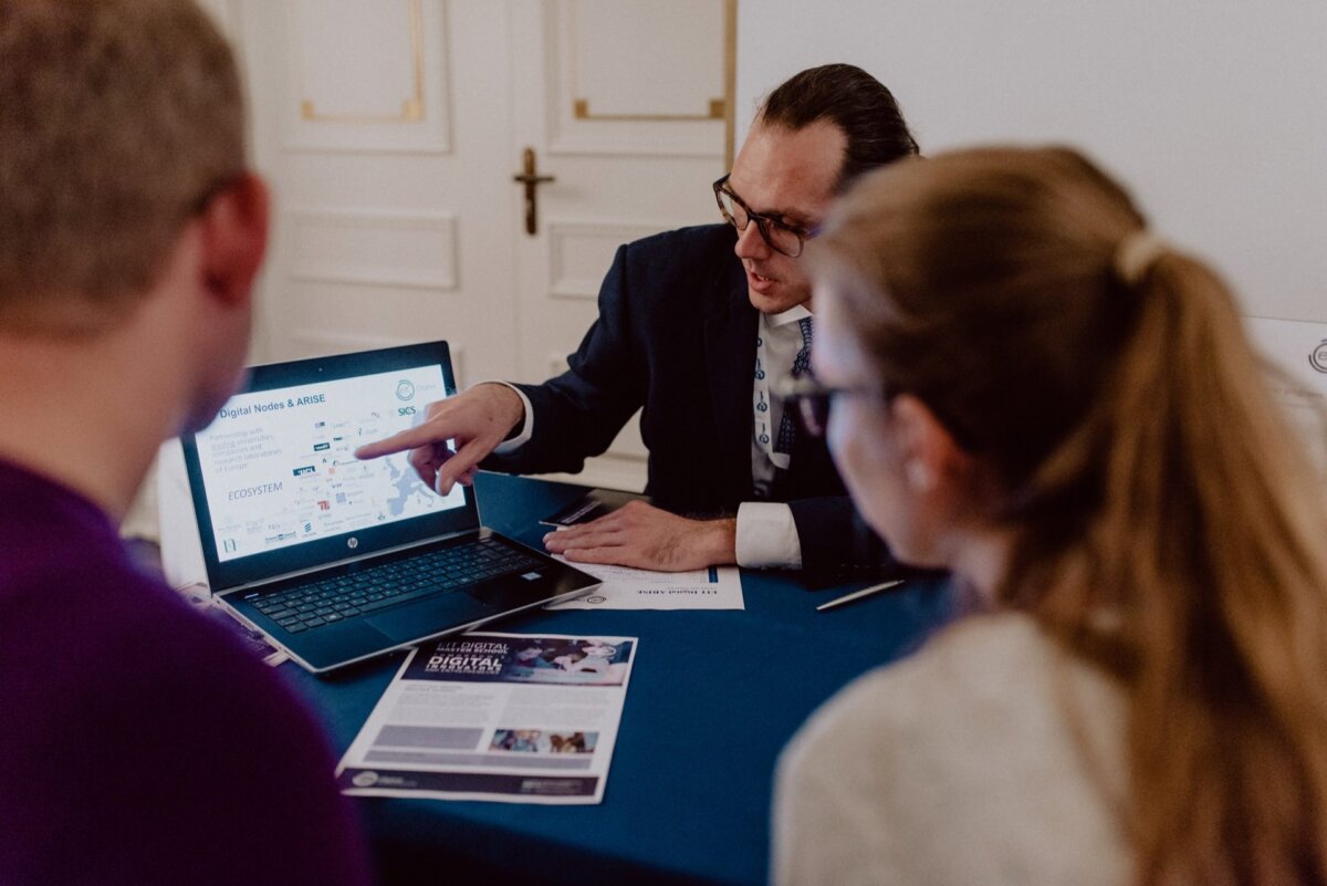 A man in glasses and a dark suit sits at a table, pointing to a laptop screen displaying various charts and graphs. Two people are listening attentively, with their backs to the camera. Papers and brochures are also spread out on the table, capturing a photo moment of the events.  