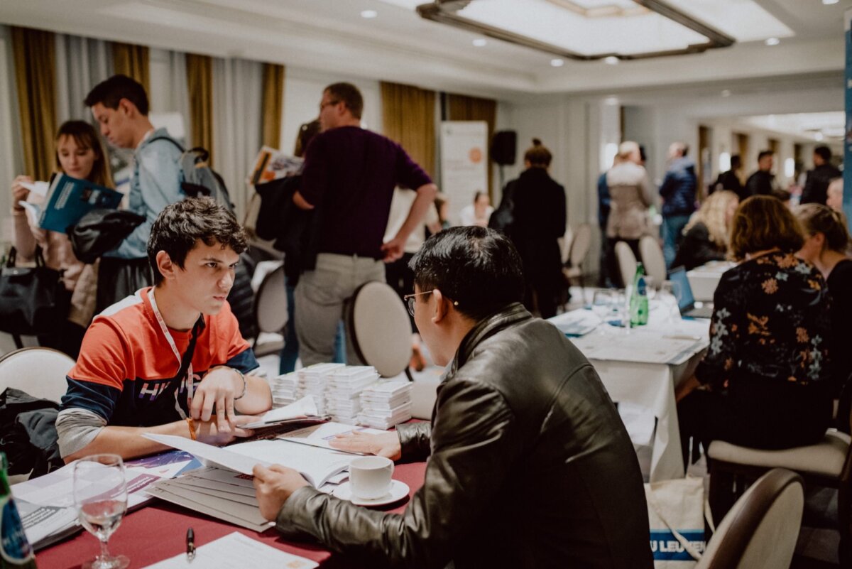 A busy conference or networking event with people talking in a well-lit room. In the foreground, two people are sitting at a table set with books, immersed in discussion. In the background, you can see more participants interacting and reviewing materials, which is ideal for event photography.  