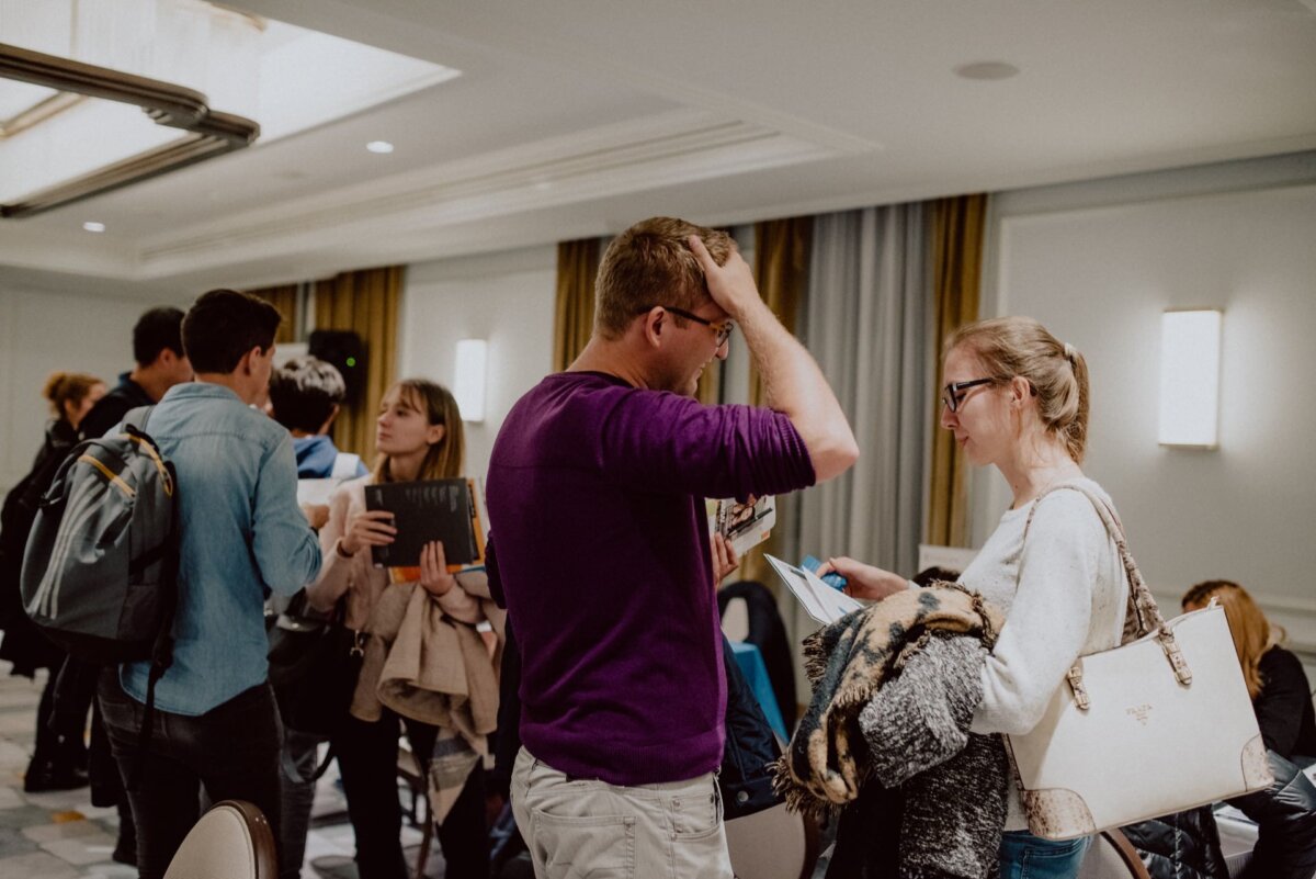 A group of people are gathered in a brightly lit conference room immersed in conversation. In the foreground, a man in a purple sweater and glasses scratches his head while talking to a woman in a cream sweater holding a book. This scene captures the essence of event photography, with others talking in the background.  