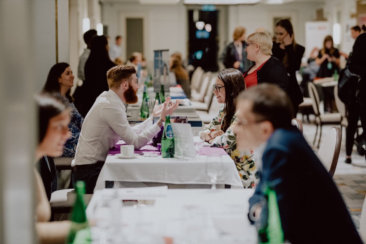 The bustling indoor stage shows people sitting in pairs at tables and immersed in discussions. It appears to be a networking or business event. There are water bottles and papers on the tables, and other people can be seen mingling in the background, which was perfectly captured by an event photographer Warsaw.  