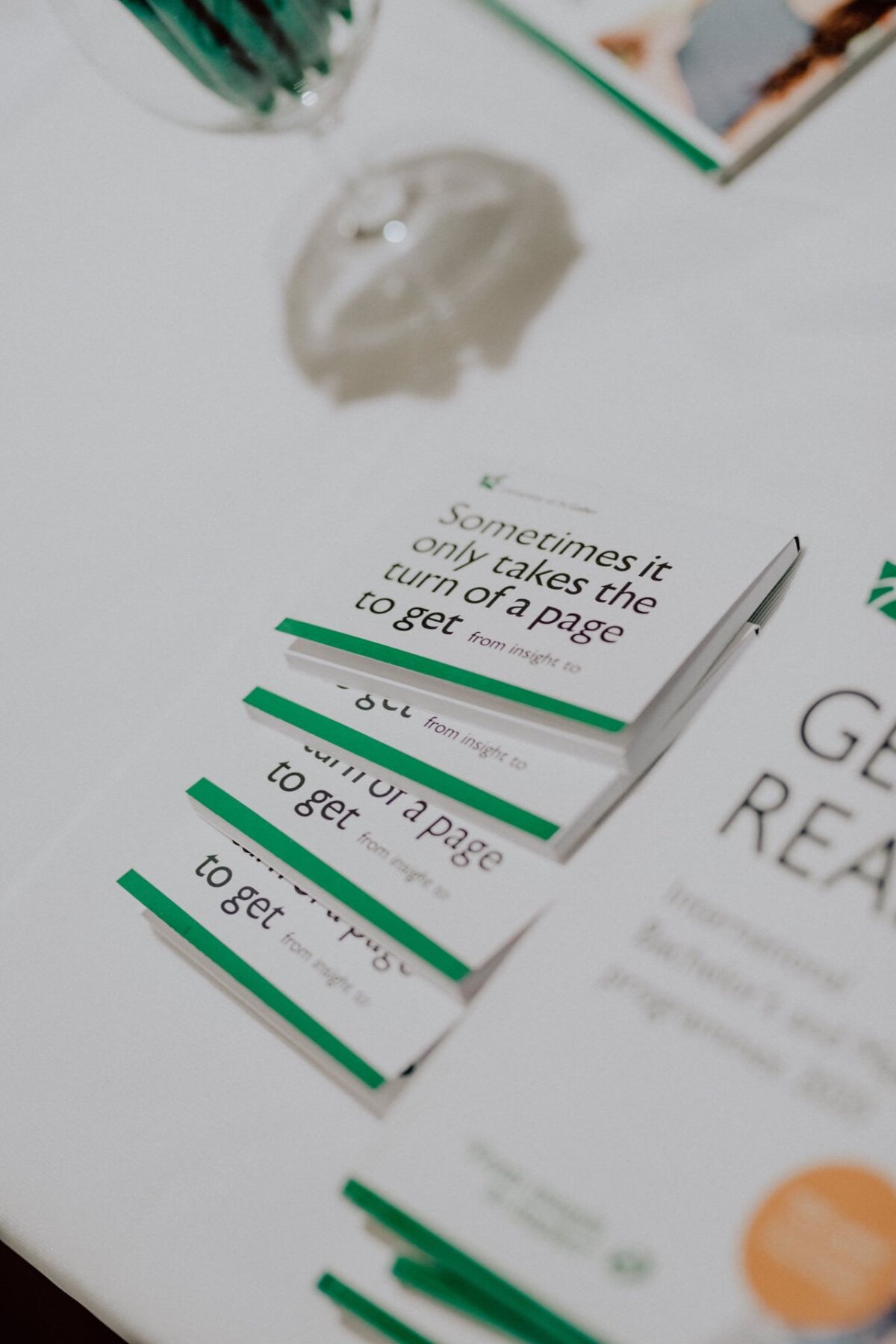 Close-up of a white table on which lie several neatly arranged books. The top book has text on the cover: "Sometimes all you have to do is turn the page to get...". Also lying on the table is part of a glass of water and another partially visible book, capturing moments reminiscent of a photo-report of events.  